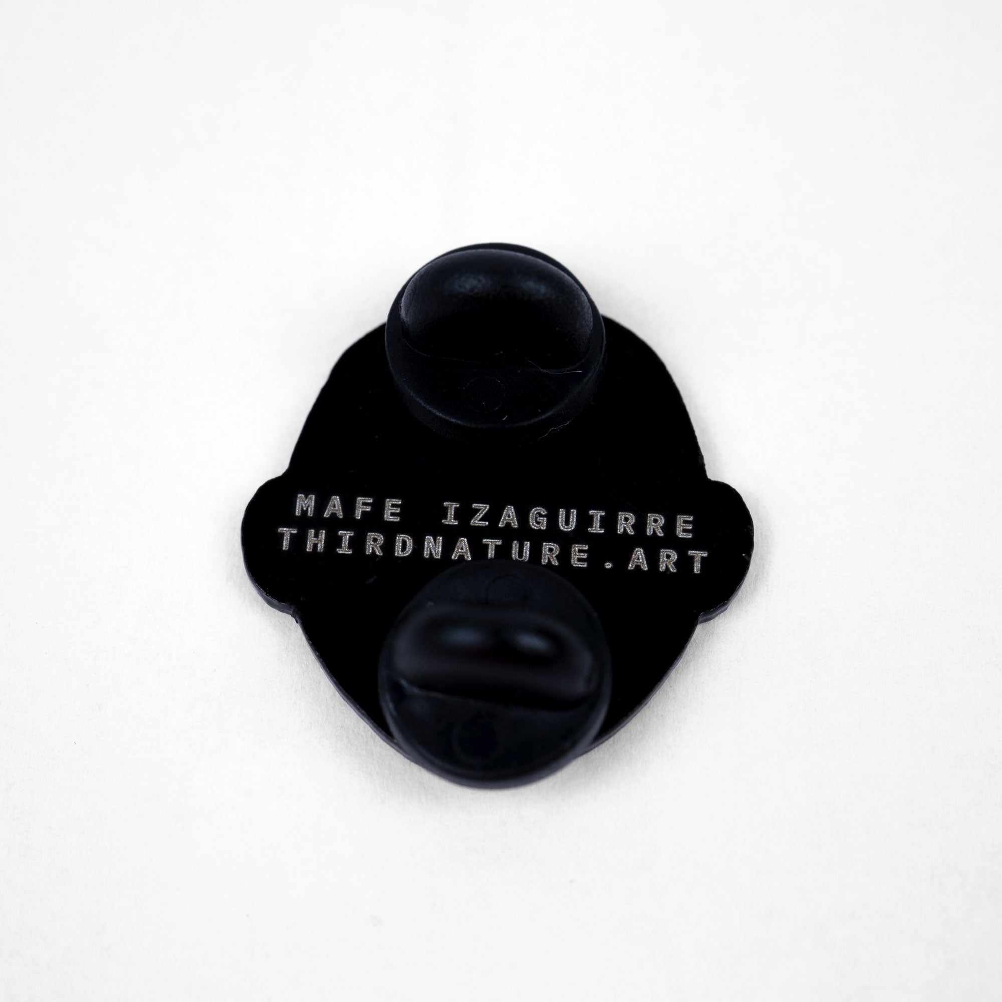 Back side of an enamel pin with text 'MAFE IZAGUIRRE THIRDNATURE.ART' on white letters