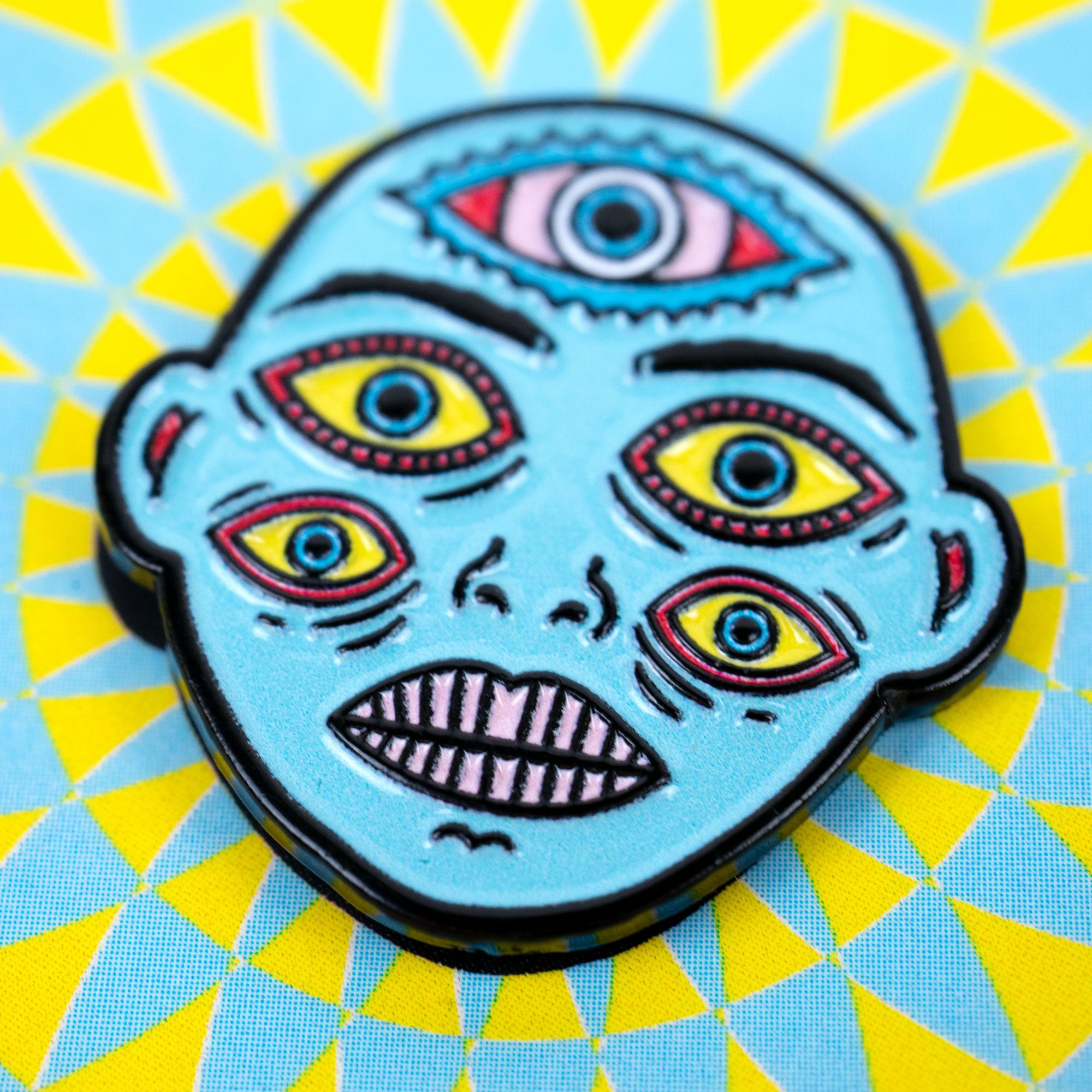 Enamel pin with a colorful face design on a geometric blue and yellow background.