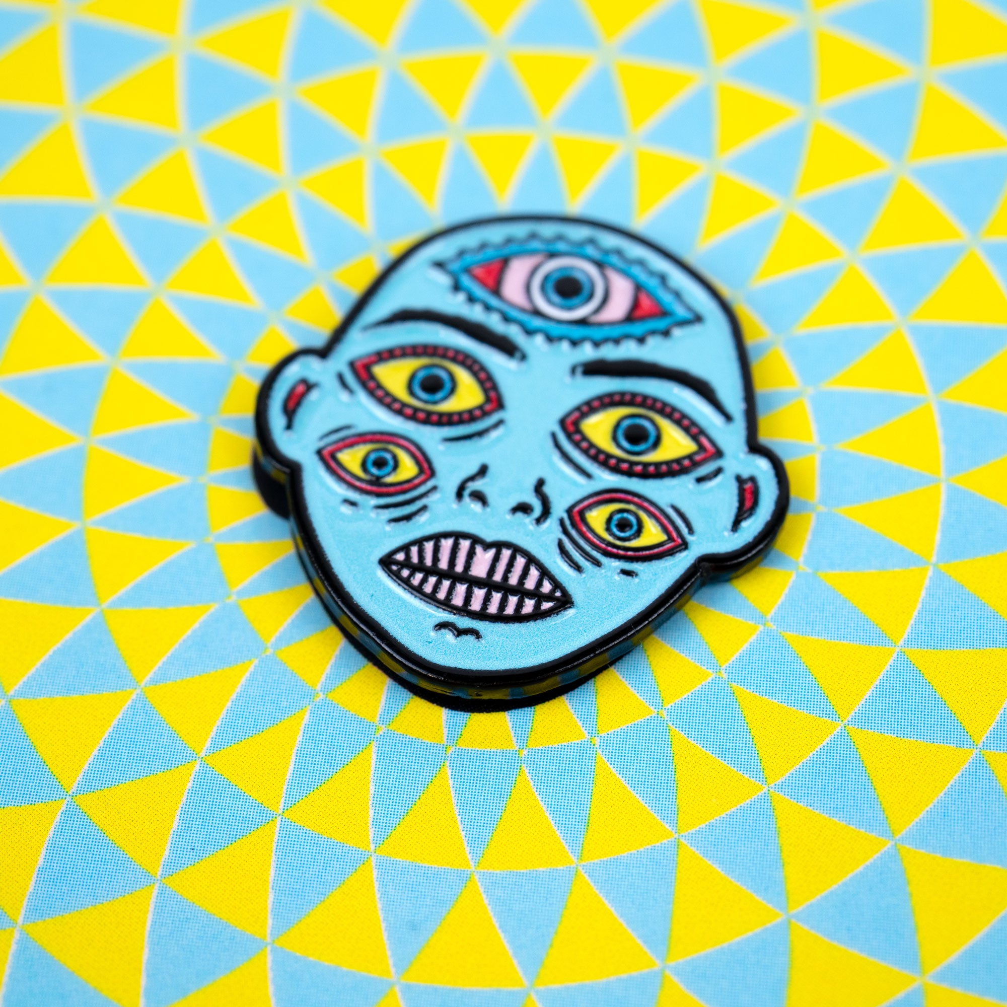 Enamel pin with a colorful face design on a geometric blue and yellow background.